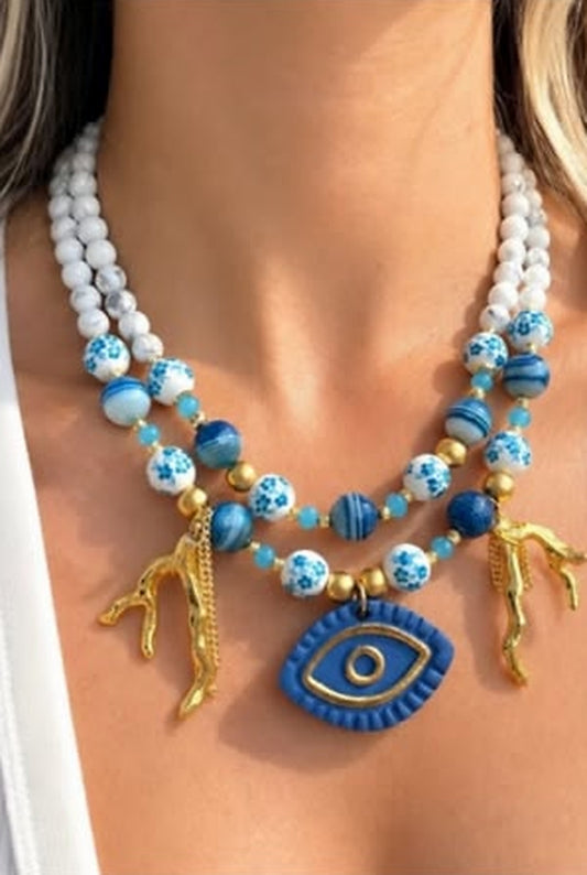 Two-strand necklace set in white and blue beads with a ceramic evil eye pendant and gold coral charms